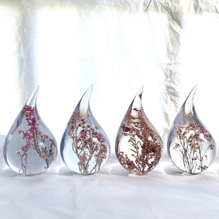 Four resin drops sculpted in a teardrop shape with purple Erica fynbos flowers embedded, positioned on a textured surface with a white background.