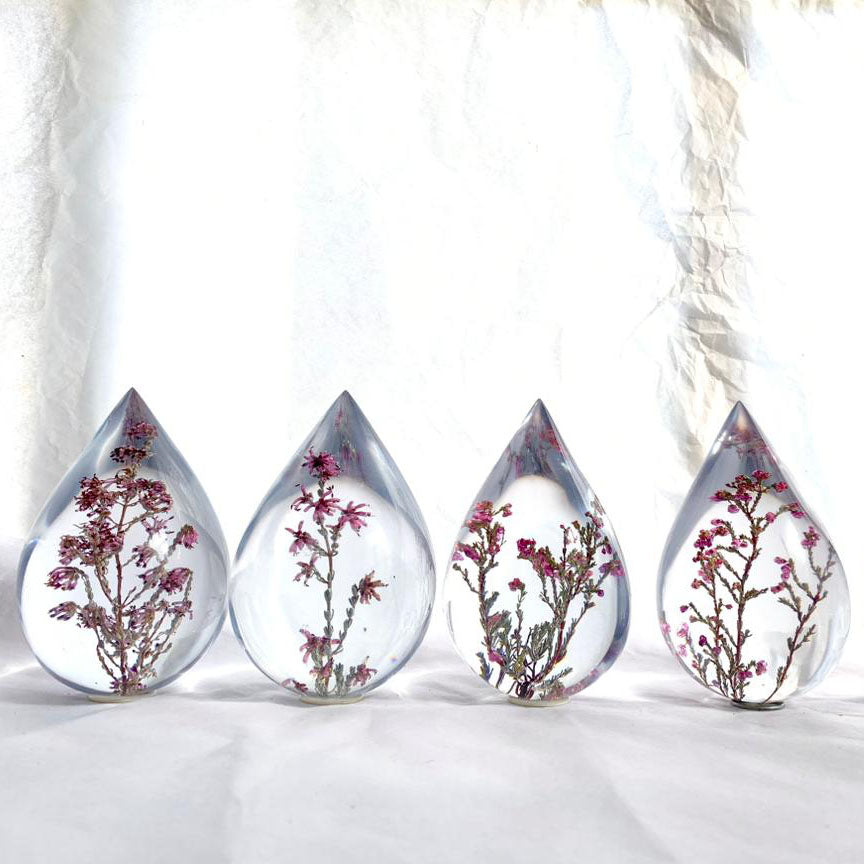 Four resin drops sculpted in a teardrop shape with purple Erica fynbos flowers embedded, positioned on a textured surface with a white background.