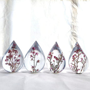 Four resin drops sculpted in a teardrop shape with purple Erica fynbos flowers embedded, positioned on a textured surface with a white background.