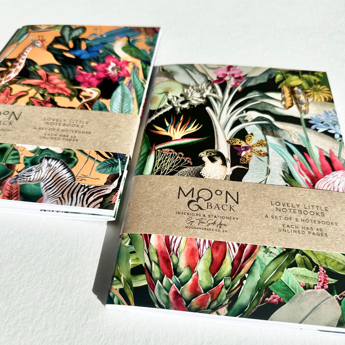 Two notebooks with colorful animal and nature-themed designs on a white surface, featuring the brand 'Moon & Back'.