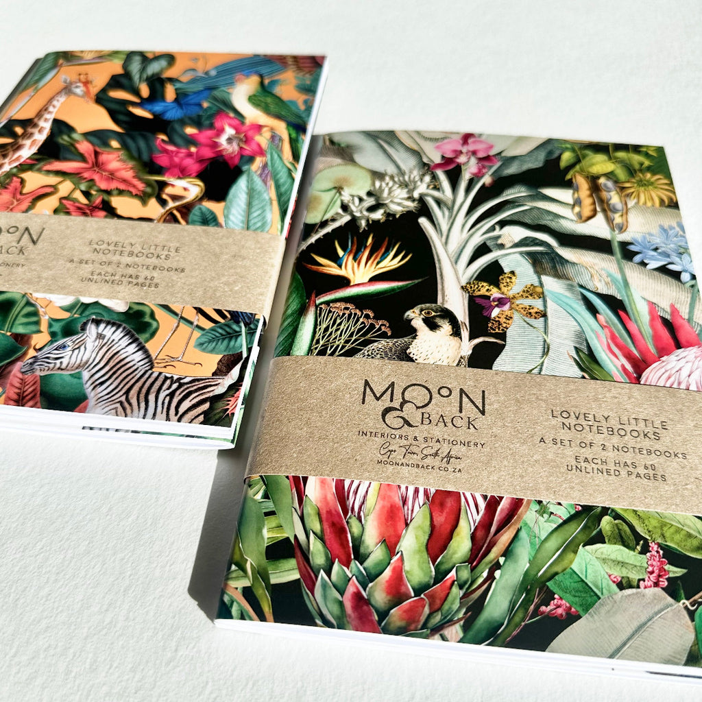 Two notebooks with colorful animal and nature-themed designs on a white surface, featuring the brand 'Moon & Back'.