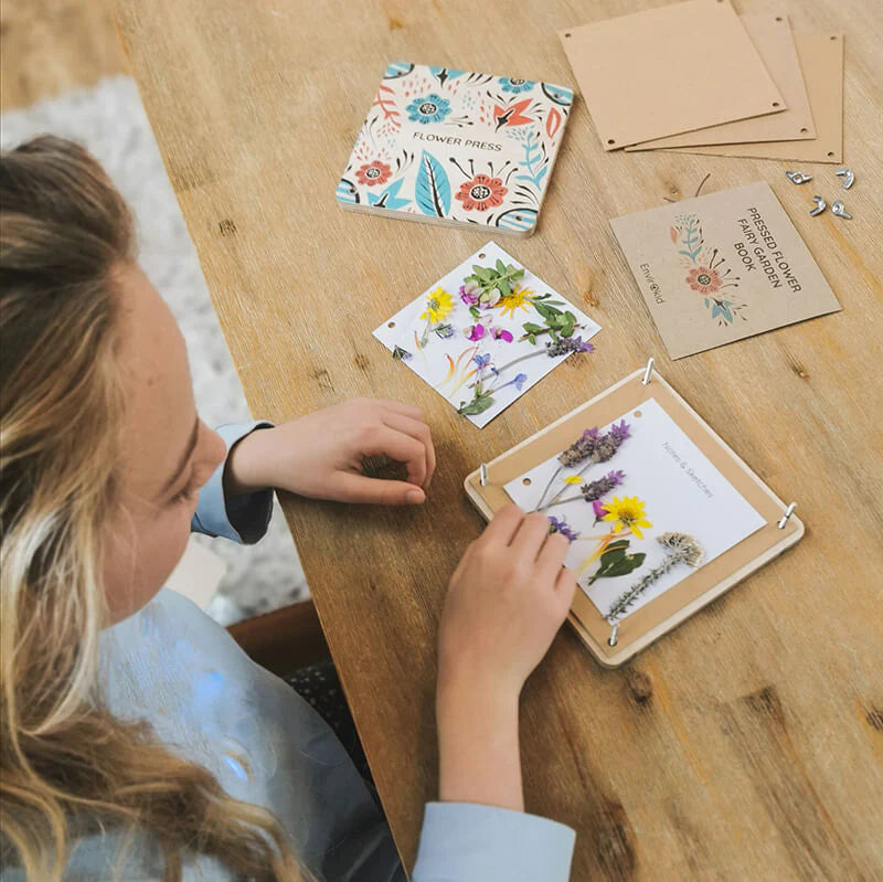 Person arranging pressed flowers on a wooden board with craft materials on a table.