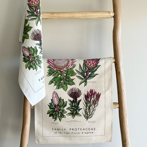Towels with botanical protea and fynbos designs hanging on a wooden ladder against a plain wall.