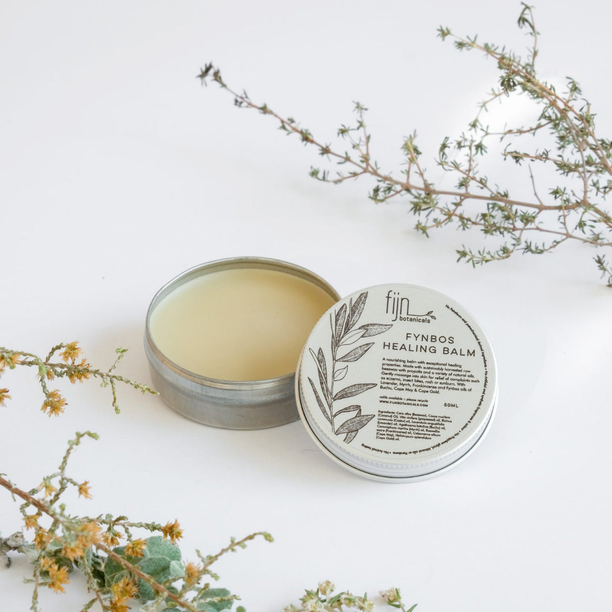 Metal tins of Fynbos balm, with the top tin open, revealing a yellowish balm inside. Surrounding the tins are sprigs of herbs and flowers, presumably some of the botanical ingredients of the product.