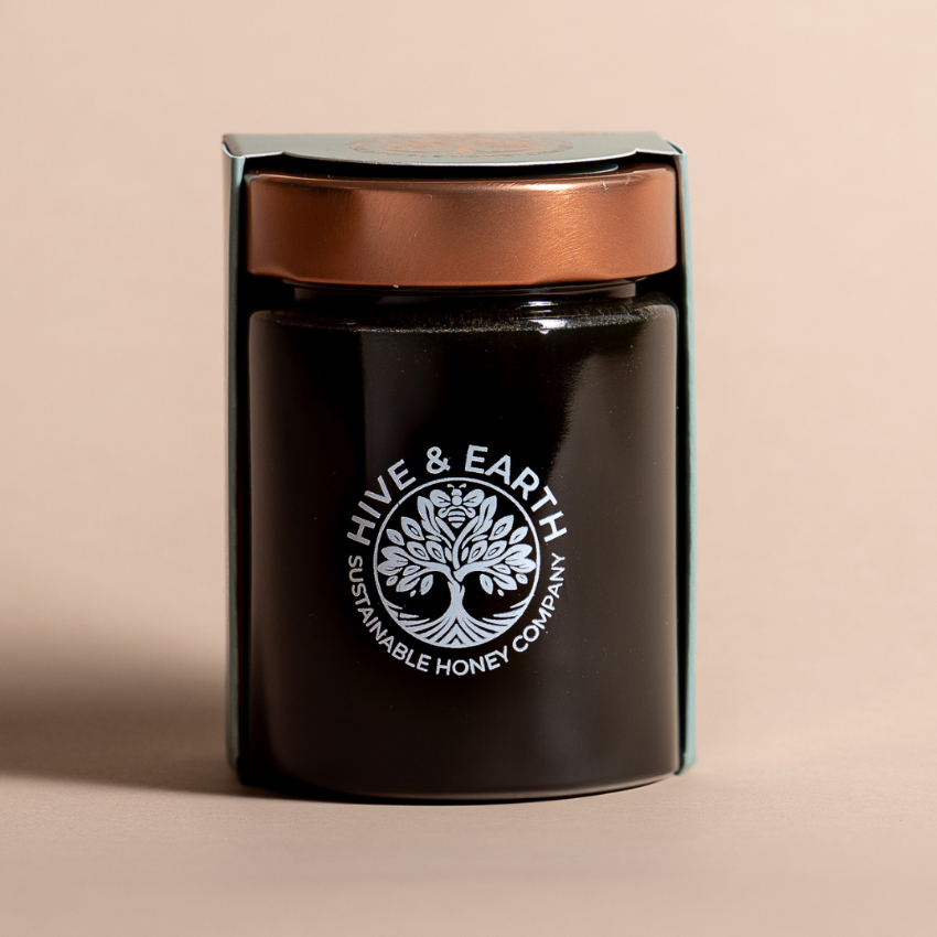 A dark glass jar of honey with a metallic copper lid, presented in its light green cardboard packaging against a solid beige background. The front of the jar features a white printed logo for 'Hive & Earth'.