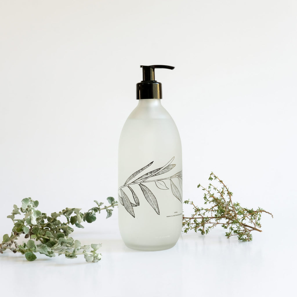 A clear glass bottle of Fynbos liquid soap with a black pump dispenser, featuring a botanical print of leaves and branches on the label, accompanied by dried botanicals.