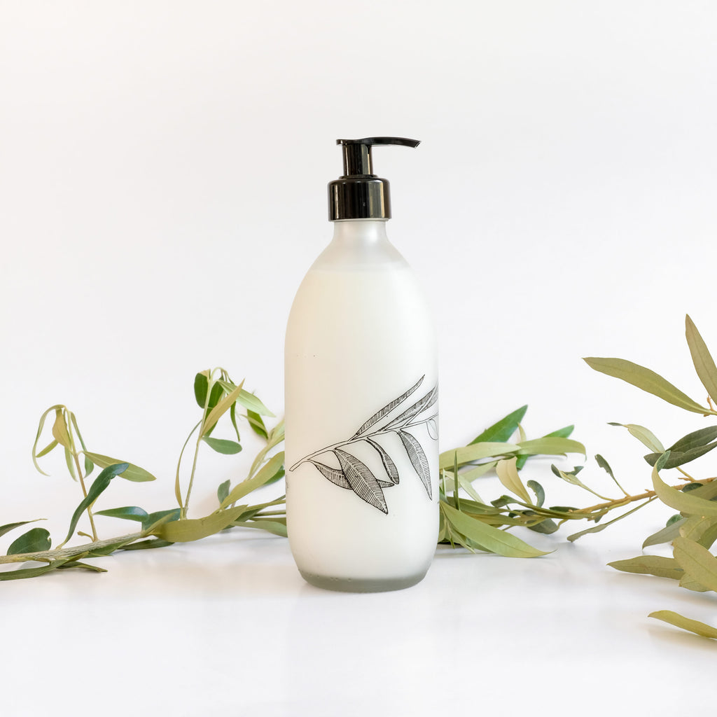 A clear glass bottle with a pump dispenser, labeled 'Fynbos body lotion', placed on a white surface with green foliage in the background.