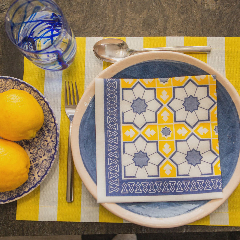 Table setting with a blue and yellow patterned napkin on a plate, lemons, and cutlery on a striped tablecloth.