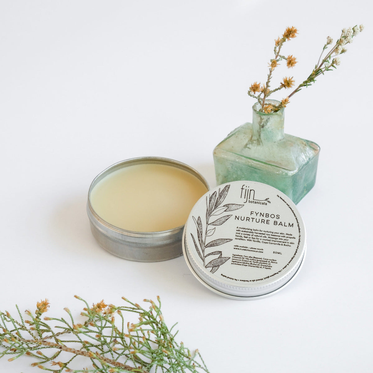 Metal tins of Fynbos balm, with the top tin open, revealing a yellowish balm inside. Surrounding the tins are sprigs of herbs and flowers, presumably some of the botanical ingredients of the product.
