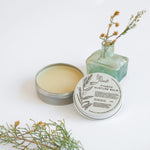 Load image into Gallery viewer, Metal tins of Fynbos balm, with the top tin open, revealing a yellowish balm inside. Surrounding the tins are sprigs of herbs and flowers, presumably some of the botanical ingredients of the product.
