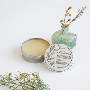 Metal tins of Fynbos balm, with the top tin open, revealing a yellowish balm inside. Surrounding the tins are sprigs of herbs and flowers, presumably some of the botanical ingredients of the product.