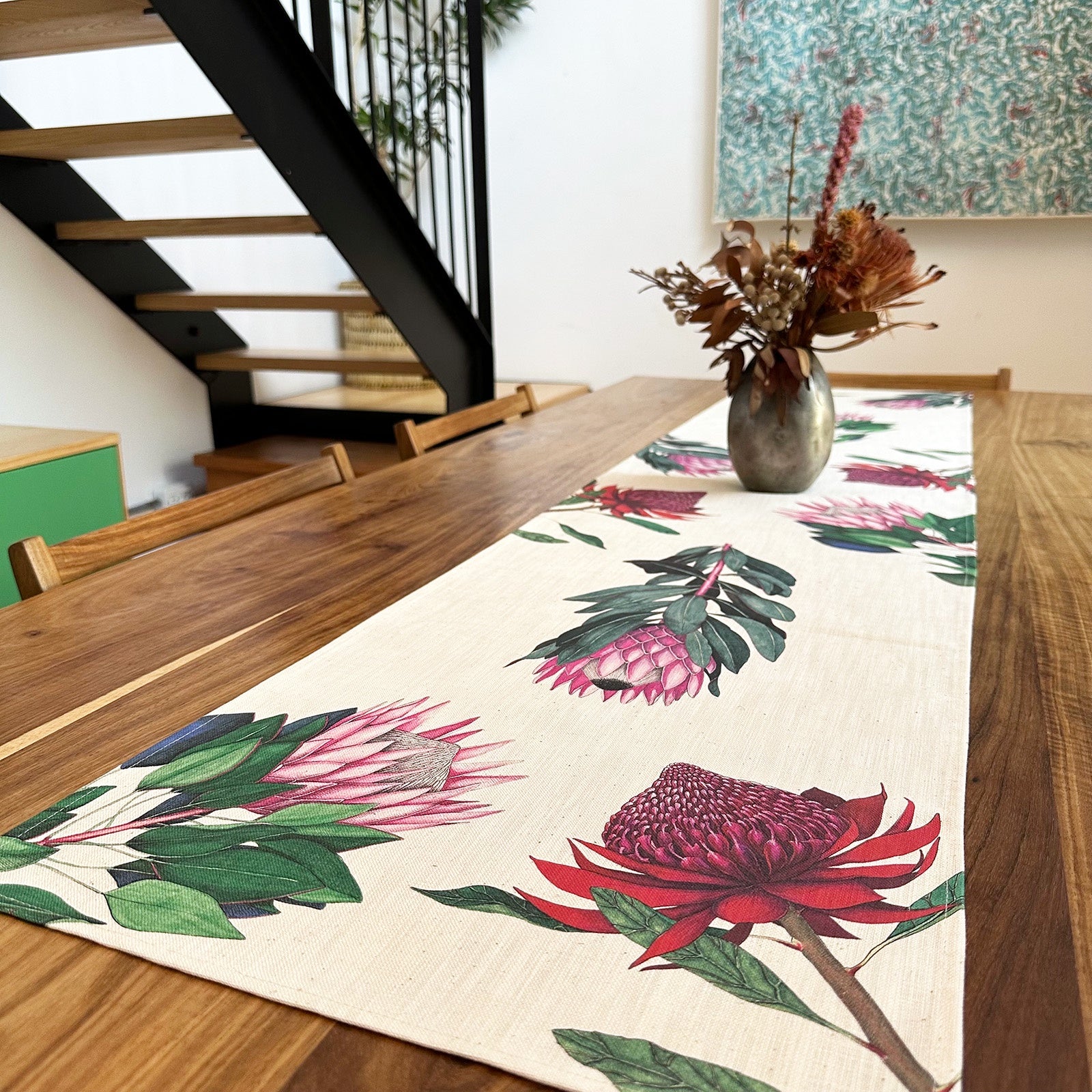 Dining table with a protea floral table runner and vase of flowers in a room with stairs and a wall art piece.