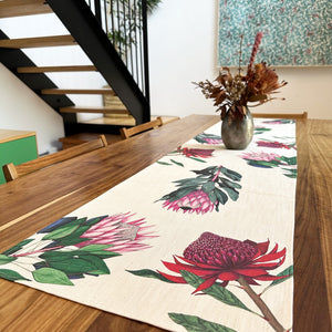 Dining table with a protea floral table runner and vase of flowers in a room with stairs and a wall art piece.