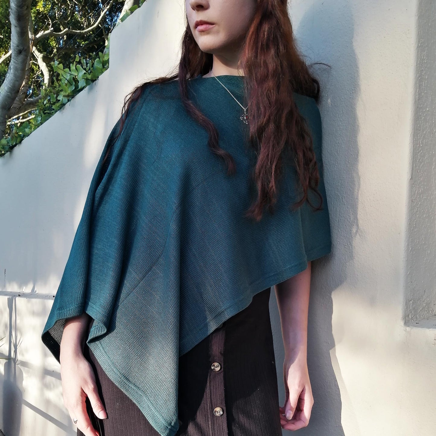 Person wearing a teal poncho against a light-colored wall with greenery in the background