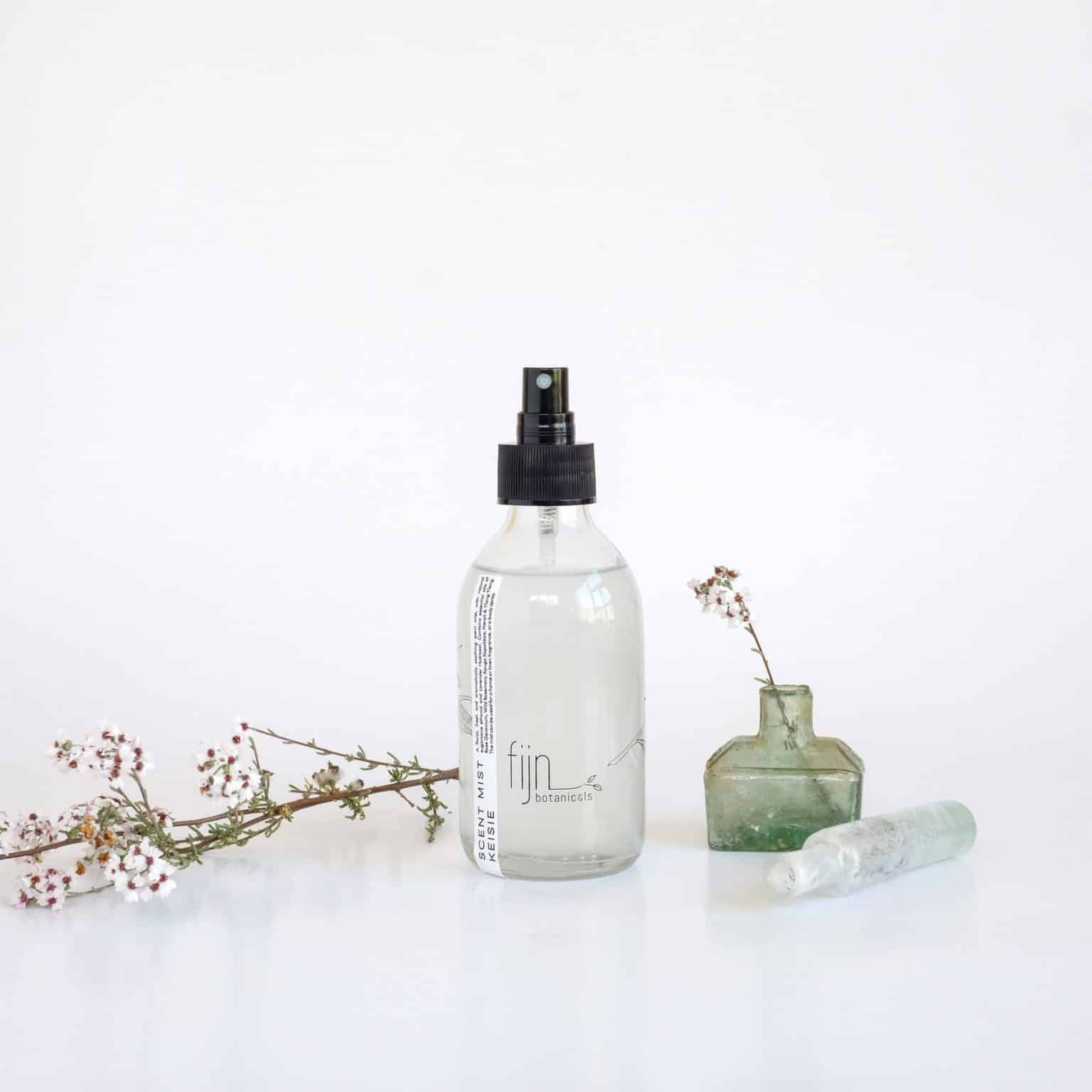 A clear glass bottle of fynbos room spray with a black spray dispenser, featuring a botanical print of leaves and branches on the label, accompanied by dried botanicals.