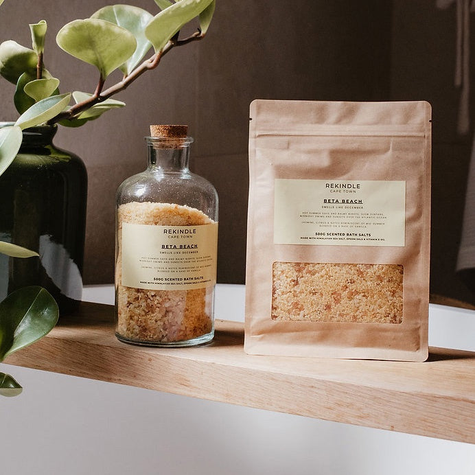 Bath salts in a glass bottle and packaging on a wooden shelf with a plant.
