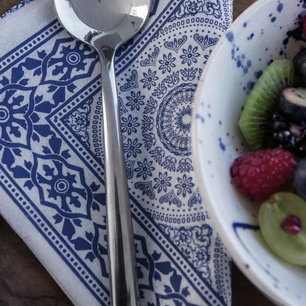 A silver spoon on a blue and white patterned napkin with a bowl of fruit in the corner.