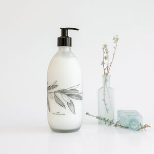 A clear glass bottle of Fynbos body lotion with a black pump dispenser, featuring a botanical print of leaves and branches on the label, accompanied by dried botanicals.
