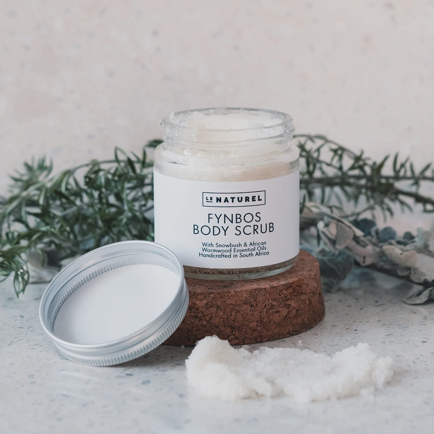 A jar of Fynbos body scrub with a lid on a stone surface, surrounded by loose scrub granules, with greenery in the background.