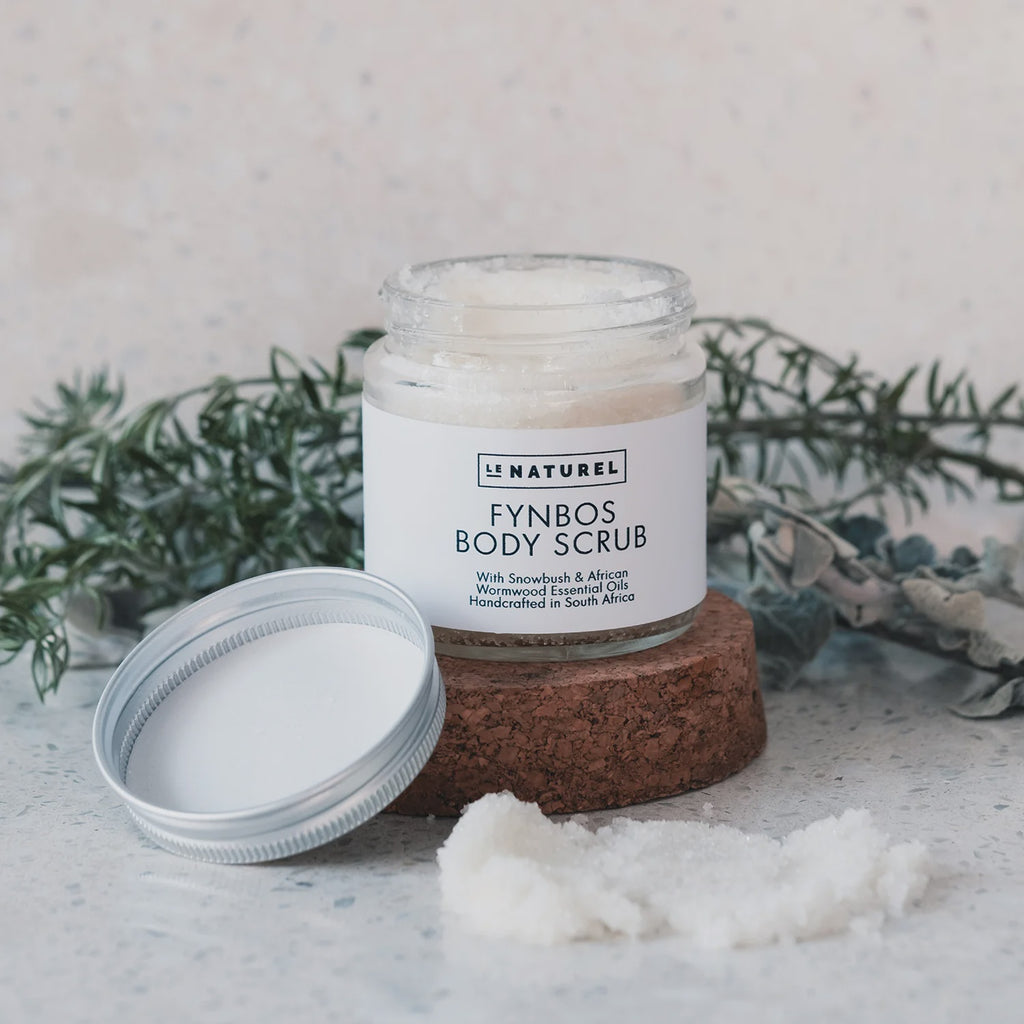 A jar of Fynbos body scrub with a lid on a stone surface, surrounded by loose scrub granules, with greenery in the background.