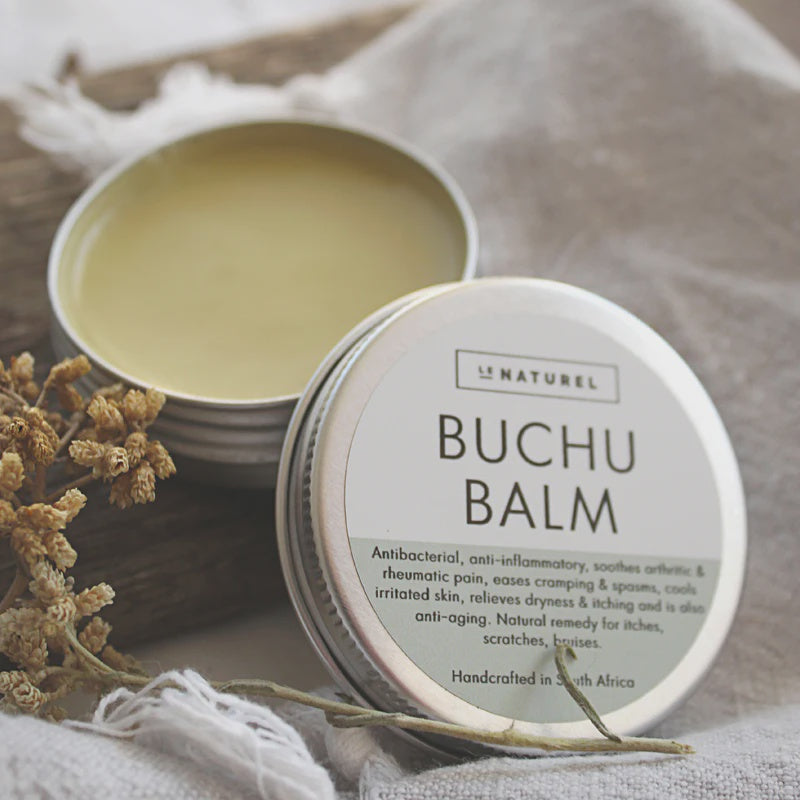 An open tin container of Buchu Balm with a label, surrounded by natural elements and a soft fabric surface.