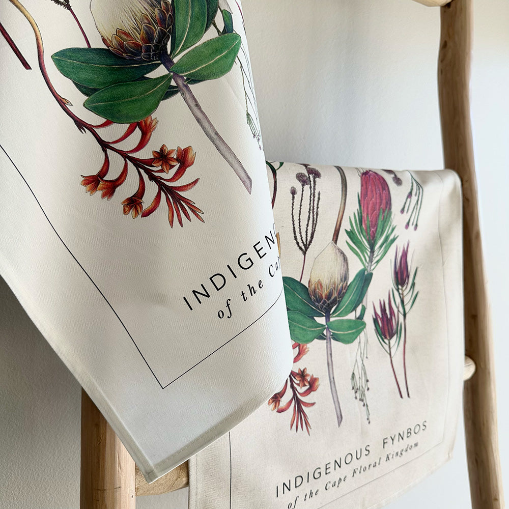 Two tea towels with botanical fynbos designs hanging on a wooden rack.