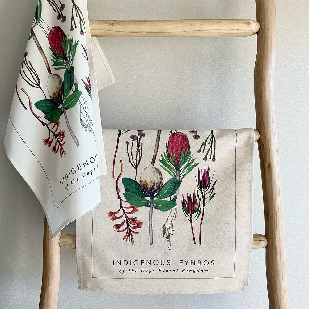 Two towels with fynbos floral designs hanging on a wooden ladder against a light gray wall.