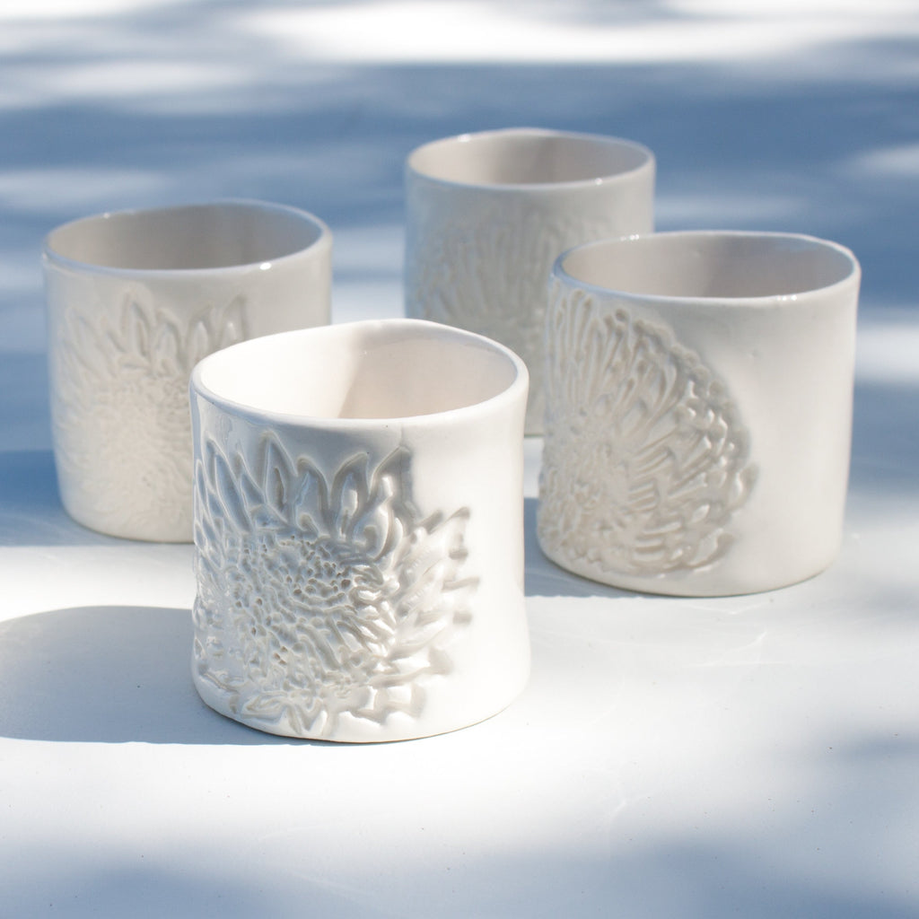 A set of four white porcelain espresso cups with embossed protea patterns, displayed on a white surface with tree shadows casting over them.