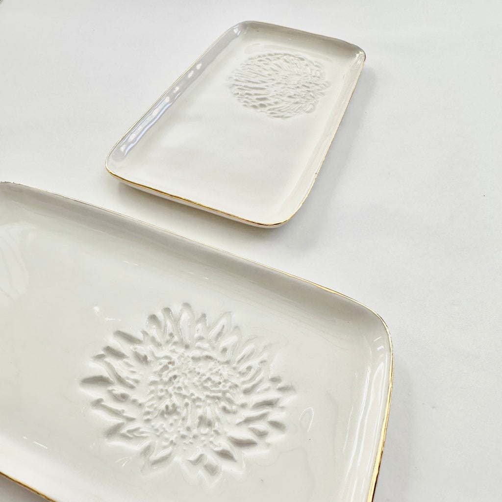 Two ceramic slates, one with a white porcelain surface and a gold rim, and the other with a king protea and pincushion protea floral pattern imprinted on it. Both are placed on a white background.