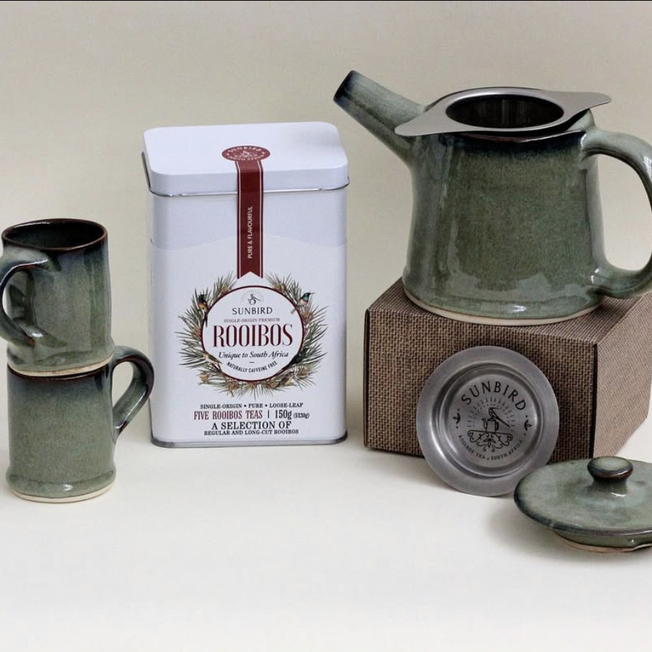 Tea set with green mugs, a teapot, and a Sunbird Rooibos tea tin on a white surface.