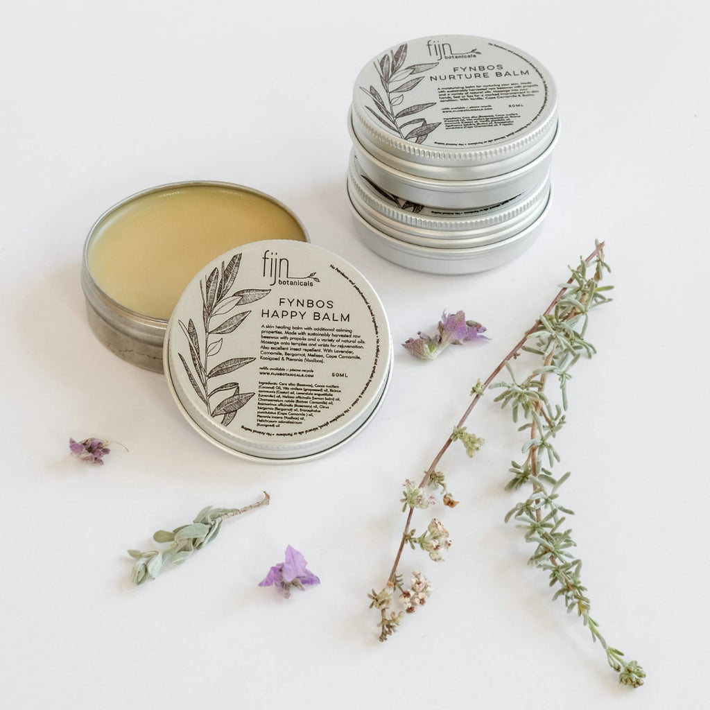 Three metal tins of Fynbos balm, with the top tin open, revealing a yellowish balm inside. Surrounding the tins are sprigs of herbs and flowers, presumably some of the botanical ingredients of the product.