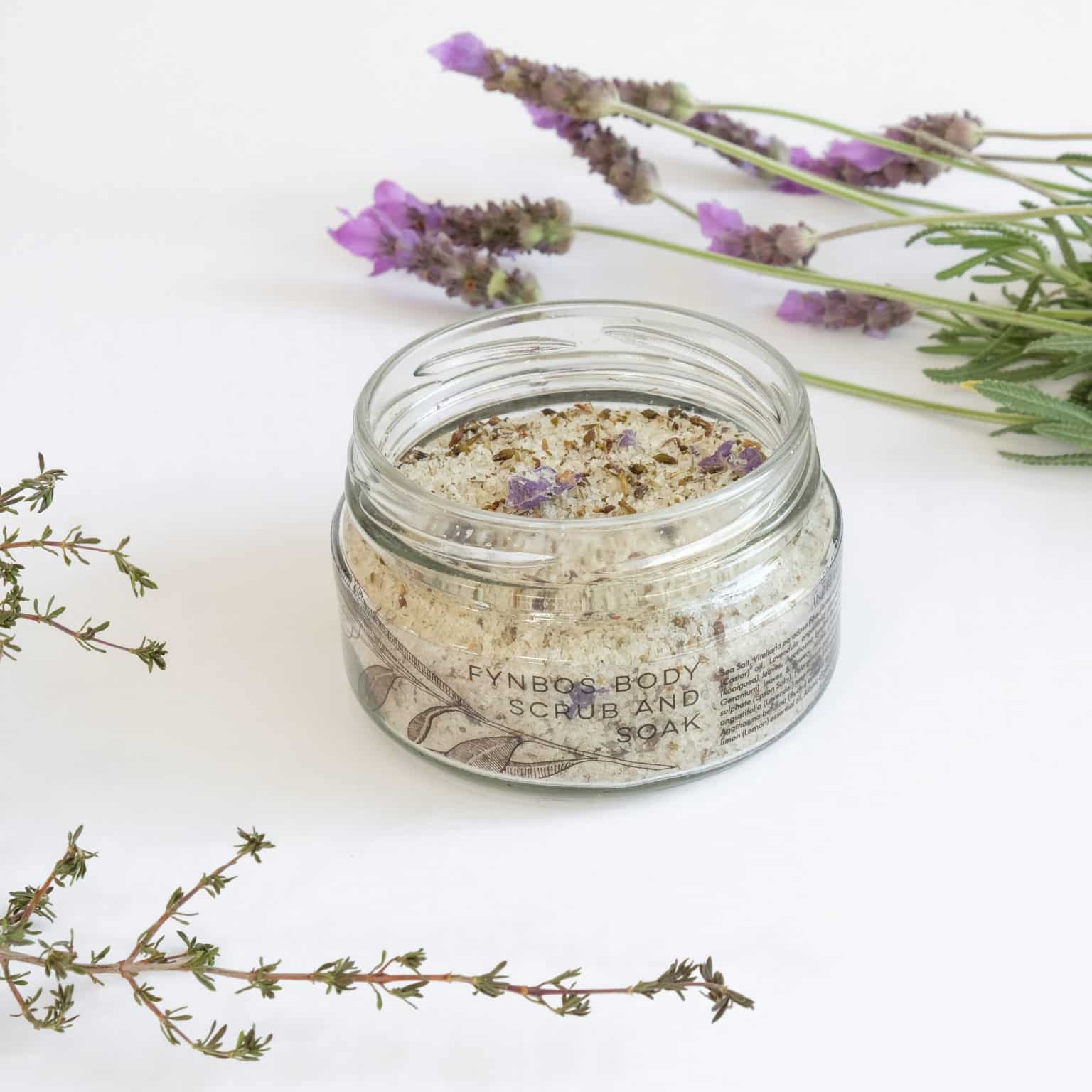 A glass jar filled with textured granules with packaging titled 'FYNBOS SCRUB AND SOAK' on a white background with lavender flowers.