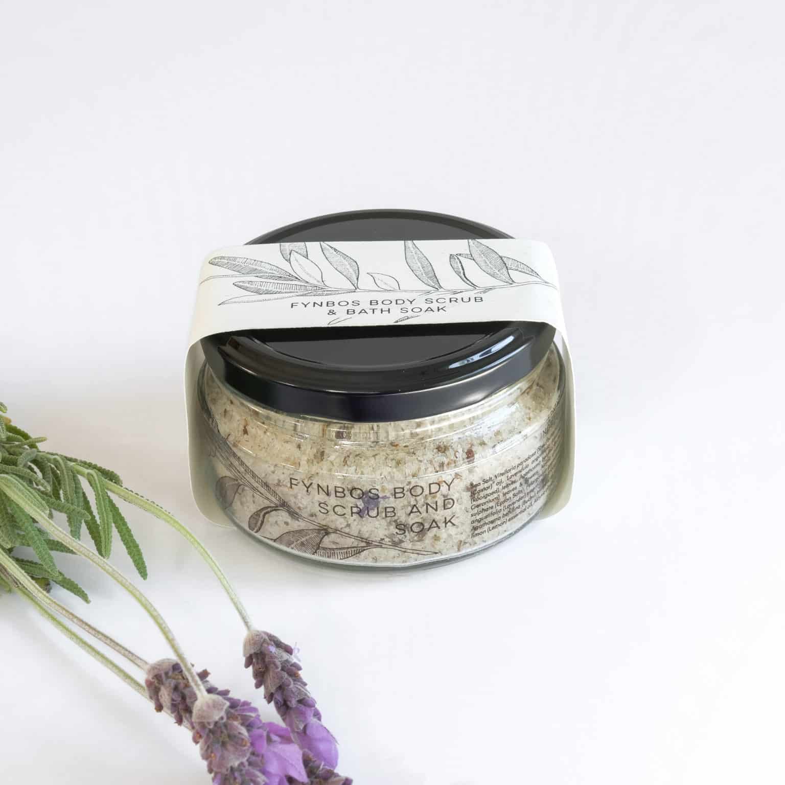 A glass jar filled with textured granules with packaging titled 'FYNBOS SCRUB AND SOAK' on a white background with lavender flowers.