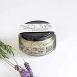 Load image into Gallery viewer, A glass jar filled with textured granules with packaging titled &#39;FYNBOS SCRUB AND SOAK&#39; on a white background with lavender flowers.
