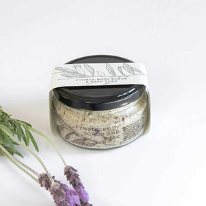 A glass jar filled with textured granules with packaging titled 'FYNBOS SCRUB AND SOAK' on a white background with lavender flowers.