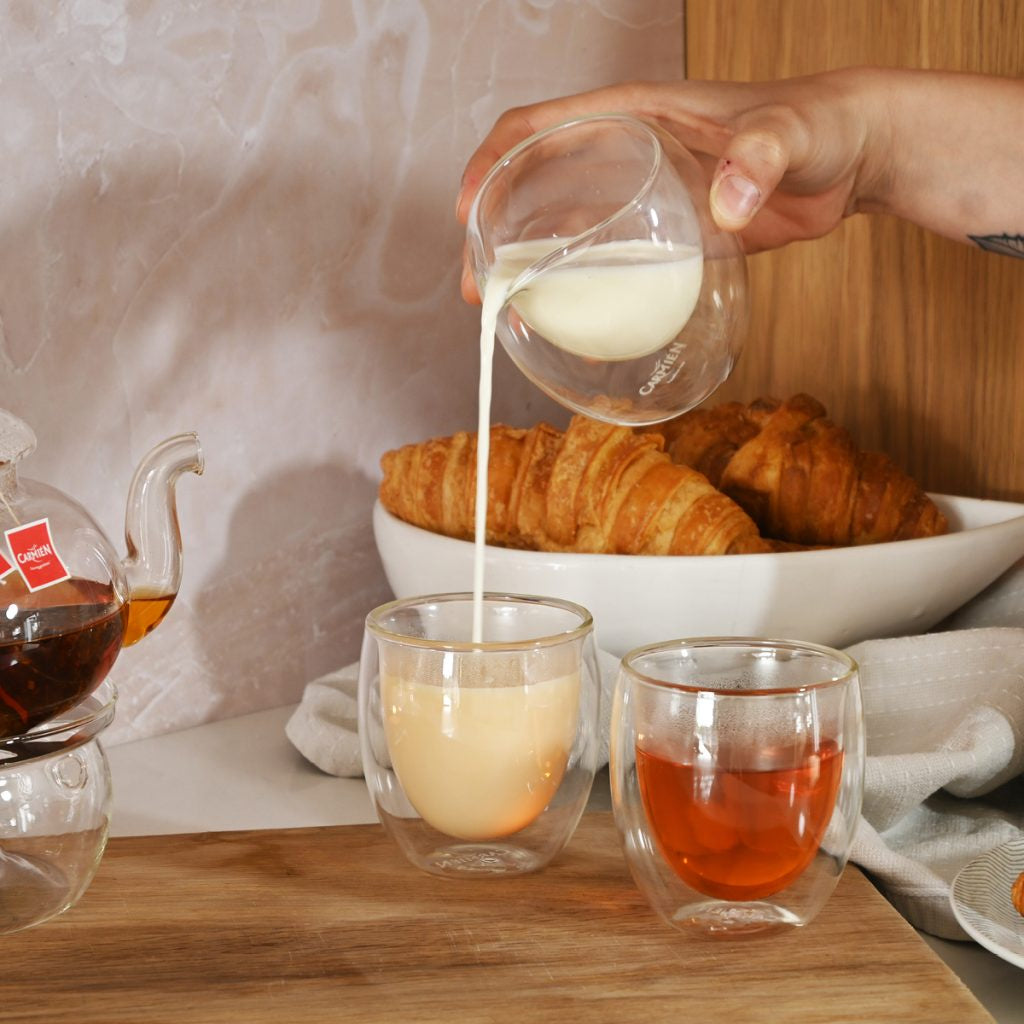 Person pouring milk into a glass on a breakfast table with croissants and coffee.
