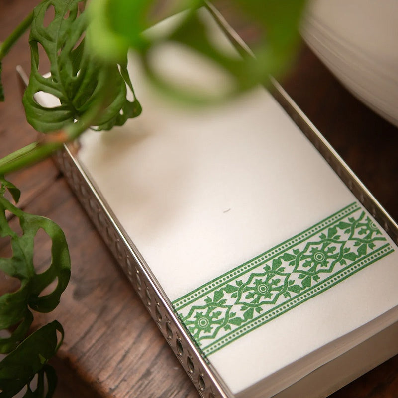 Serviette holder filled with white napkins with green decorative border on wooden surface with plant.