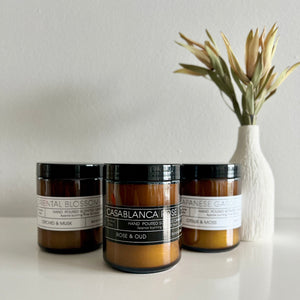 Three candles with labels on a white surface, with a vase of dried plants in the background.