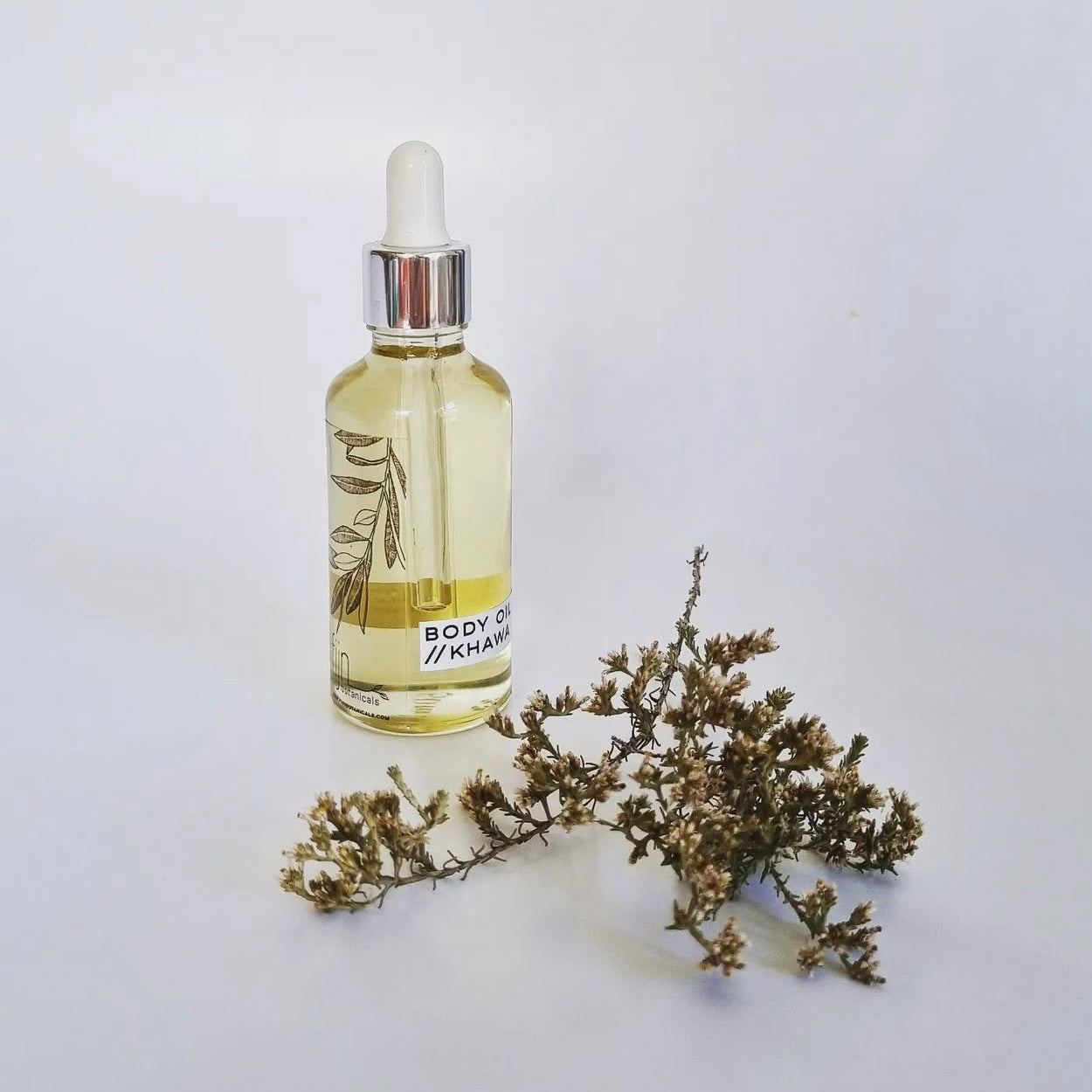 A clear glass bottle with a white dropper filled with light yellow body oil, labeled 'BODY OIL KHAWA'