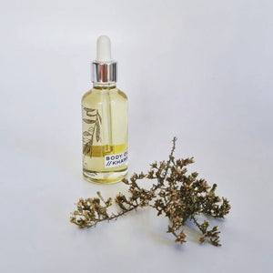A clear glass bottle with a white dropper filled with light yellow body oil, labeled 'BODY OIL KHAWA'