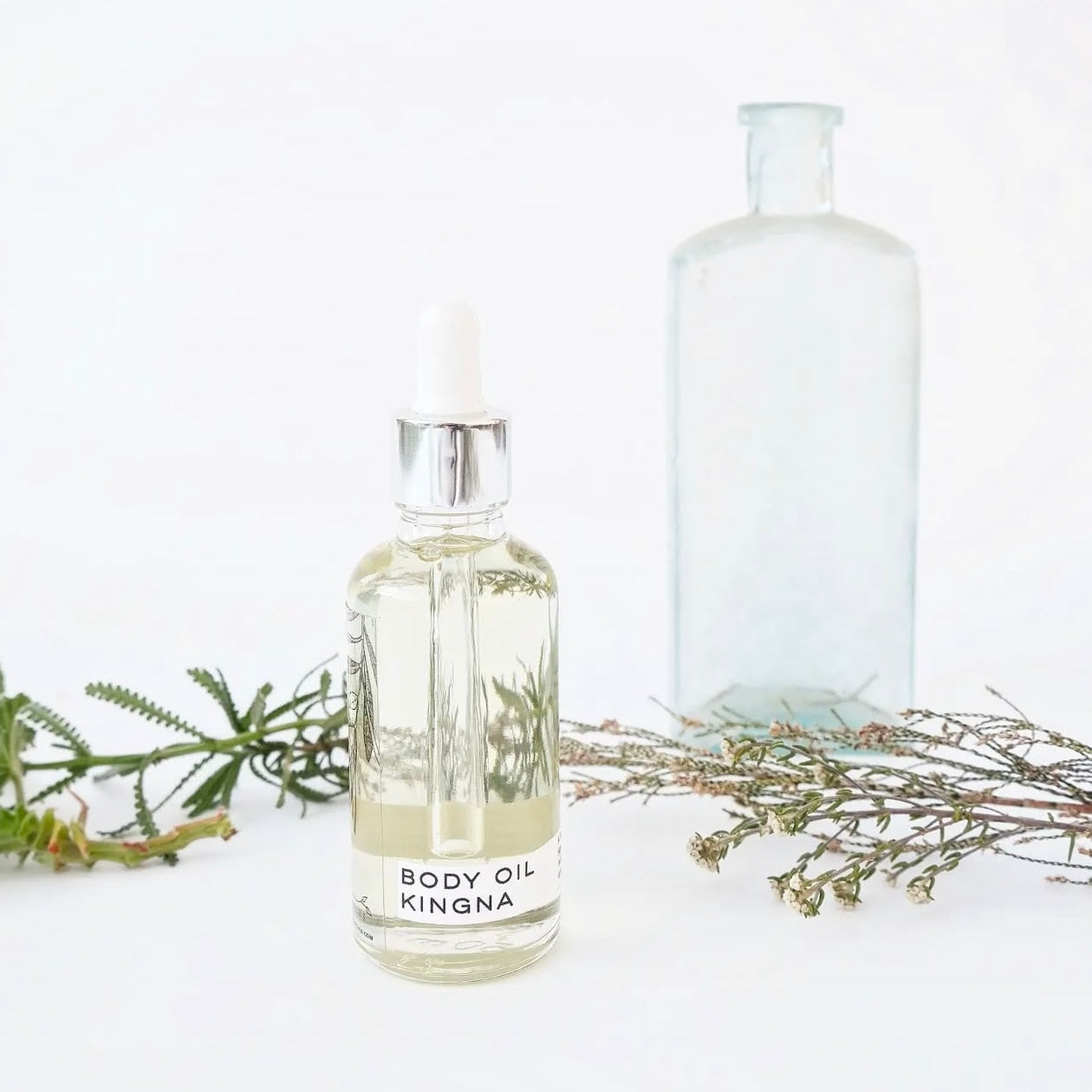 A clear glass bottle with a white dropper filled with light yellow body oil, labeled 'BODY OIL KINGNA', alongside a transparent bottle and natural elements.