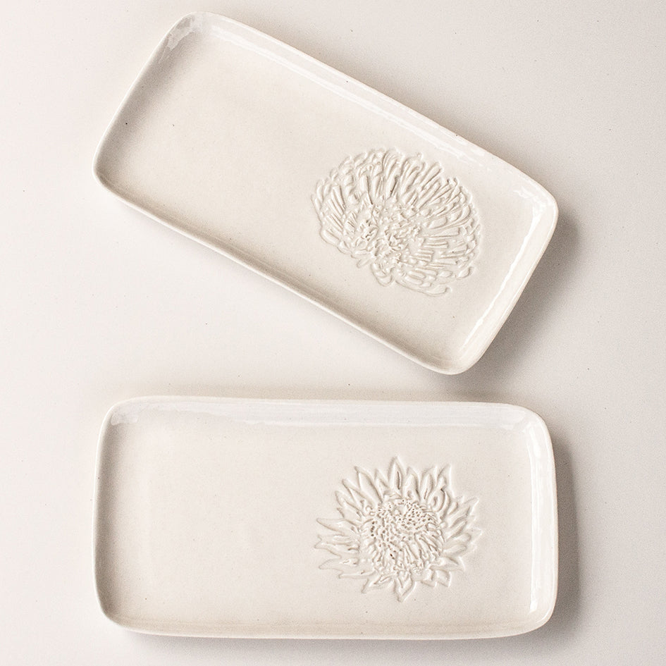 Two white ceramic slates, one with with a pincushion protea and the other with a king protea embossed on the right hand side. 