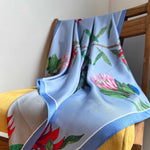 Load image into Gallery viewer, A close-up image of a reversible silk scarf with colourful protea illustrations draped on a chair. 
