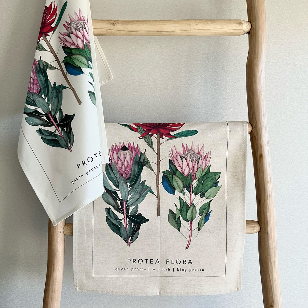 Two towels with protea flower designs hanging on a wooden ladder against a light gray wall.
