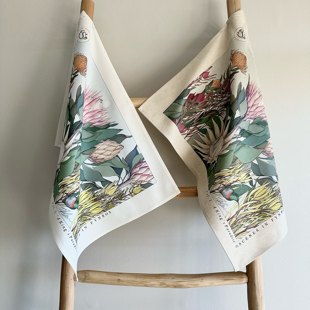 Two fynbos and protea floral-patterned towels hanging on a wooden rack against a plain background