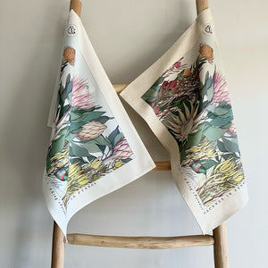 Two fynbos and protea floral-patterned towels hanging on a wooden rack against a plain background