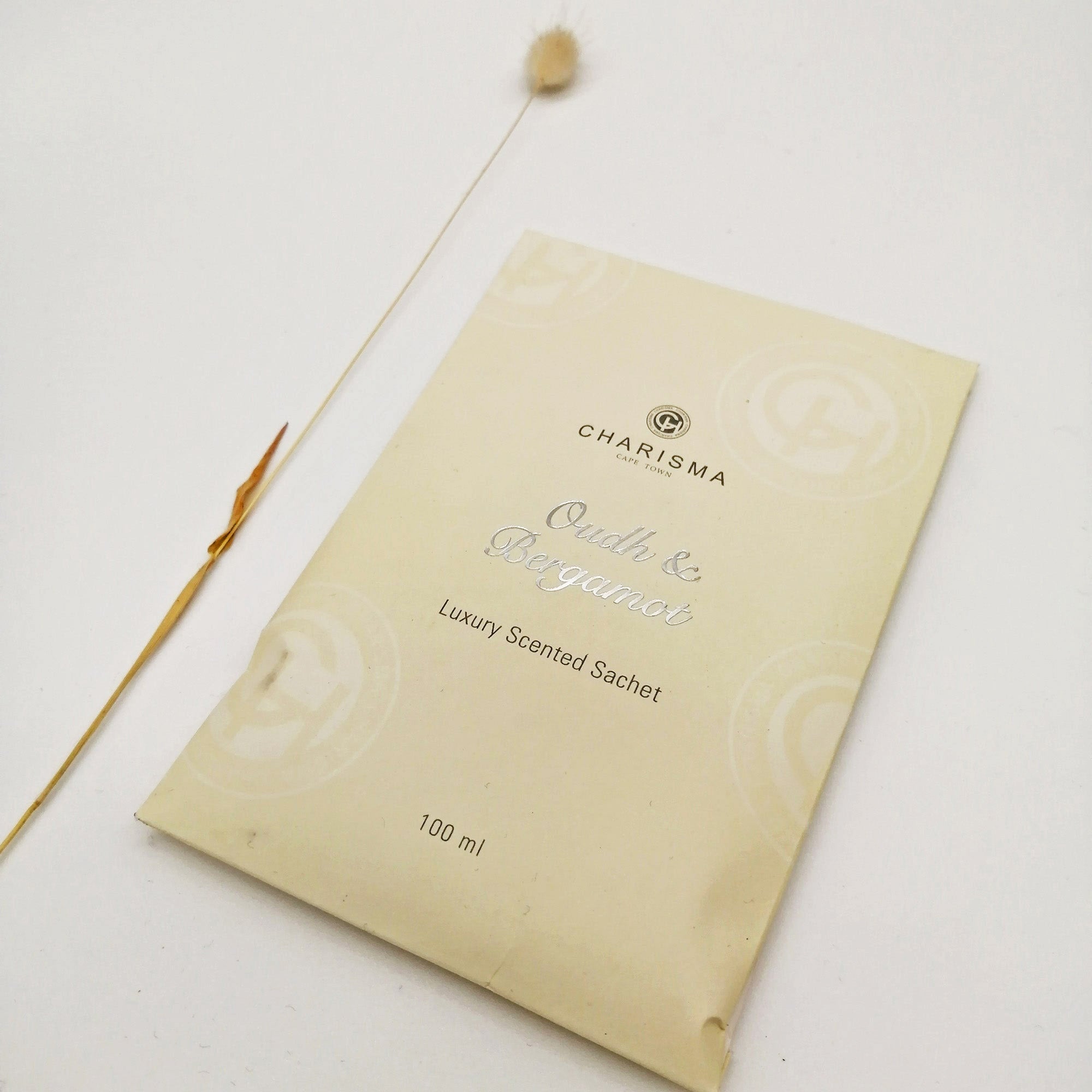 A cream coloured sachet with the title 'OUDH & BERGAMOT LUXURY SCENTED SACHET' in silver foiling alongside a dried grass.