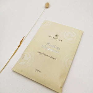 A cream coloured sachet with the title 'OUDH & BERGAMOT LUXURY SCENTED SACHET' in silver foiling alongside a dried grass.