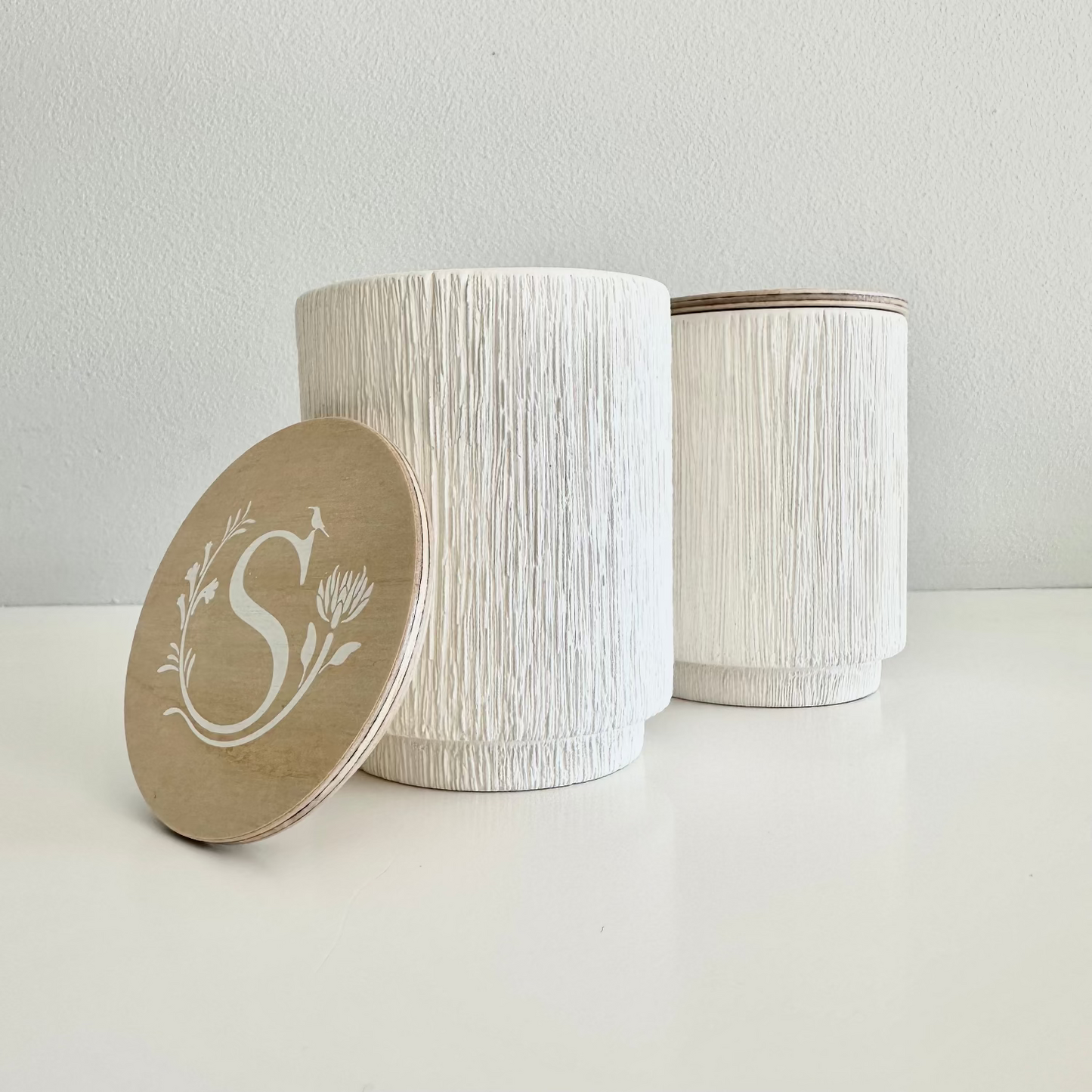 Two white textured jars with a lid featuring a monogram on a light gray background