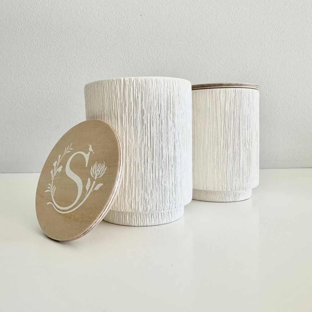 Two white textured jars with a lid featuring a monogram on a light gray background